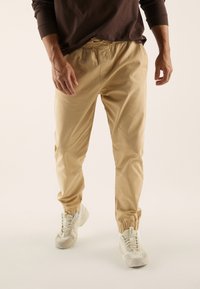 Beżowe spodnie dresowe z elastycznym pasem i regulowanym sznurkiem, zwężanymi nogawkami oraz elastycznymi ściągaczami, zestawione z jasnymi sneakersami.