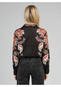 Blouse noire avec des motifs floraux multicolores, comporte un col, des fermetures à boutons et des manches longues. Portée avec un jean gris foncé.
