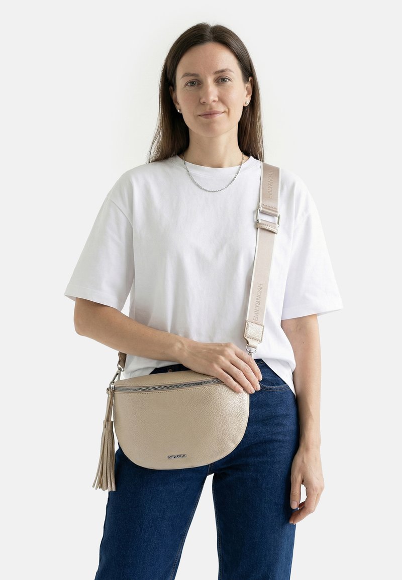 Woman with long brown hair wearing a white t-shirt and blue jeans, holding a beige crossbody bag with a tassel and wide adjustable strap.