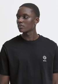 Tricou negru din bumbac cu mâneci scurte, având un mic grafic alb reprezentând un soare și textul "calm." brodat pe partea stângă a pieptului.