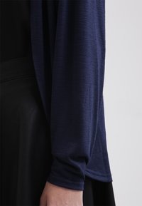 Námořnická modrá halenka s dlouhým rukávem, vyrobená z texturované látky, s volnějším střihem a širokým manžetou. Spodní část je z černého materiálu.