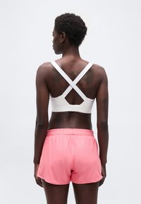 Γυναίκα που στέκεται με την πλάτη γυρισμένη, φοράει λευκό αθλητικό σουτιέν με χιαστί λουριά και ροζ αθλητικά σορτς Under Armour, μπροστά σε απλό φόντο.