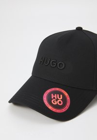 Svart baseballcap med preget "HUGO"-logo foran. Har et rundt merke med flerfarget merkevare. Stoffet har en jevn tekstur.