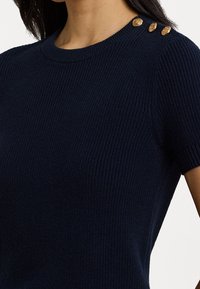 Tummansininen lyhythihainen villapaita, jossa on ribatuttu pinta, ja vasemmassa olkapäässä kolme kullanväristä koristepainiketta.