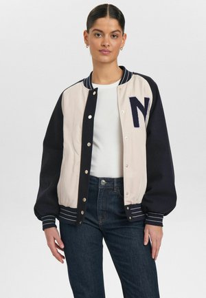 Cream and navy varsity jacket with large "N" patch, snap buttons, striped ribbed cuffs, and a relaxed fit. Made of textured fabric.