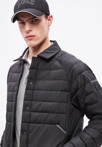 Fekete steppelt dzseki, gombos elülső résszel, steppelt mintával és oldalzsebbel. Szürke galléros pólóval és fekete sapkával viselve.