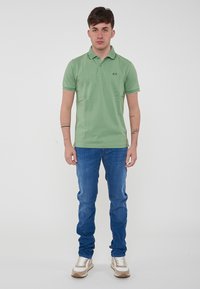 Polo verde con colletto e maniche corte, abbinato a jeans blu e sneakers beige, presenta un piccolo logo sul petto.