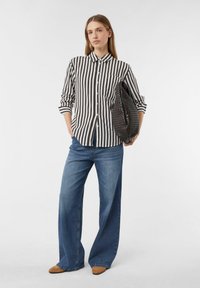 Schwarz-weißer gestreifter Hemd, blaue weite Jeans und braune Ballerinas. Das Hemd hat eine Tasche und aufgerollte Ärmel. Eine geflochtene Handtasche ist ebenfalls enthalten.