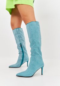Botine din material texturat, de culoare turcoaz strălucitor, până la genunchi, cu vârf ascuțit și detaliu cu fermoar pe lateral. Prezintă un toc înalt, de tip stiletto.