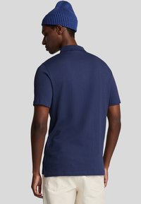 Marineblå kortærmet polo-shirt med tekstureret stof, der har en flad krave og diskrete sideslids. Bæret med off-white bukser.