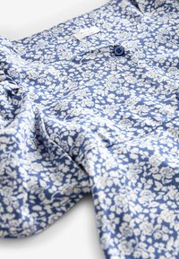 Tissu à motifs floraux bleus avec des fleurs blanches, présentant un détail de bouton et une étiquette de marque, mettant en avant une finition douce et texturée.