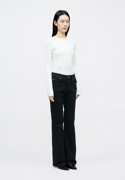 Haut blanc à manches longues avec des détails de boutons sur le côté, associé à un jean noir évasé taille haute, avec poches et accents rouges subtils.