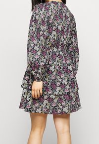 Kvinna som bär en långärmad, lager-på-lager blommig klänning med lila, vita och blå blommor mot en svart bakgrund.