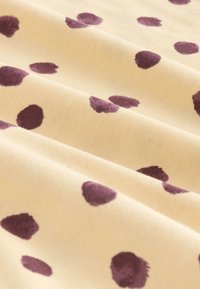 Tissu beige à pois violets foncés irréguliers, présenté avec des plis doux qui mettent en valeur la texture et le motif.