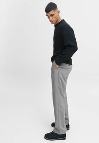 Pulover negru tricotat cu mansete cu dungi, pantaloni gri tailored și pantofi din velur negru. Modelul stă de profil cu mâinile în buzunare, fundal minimalist.