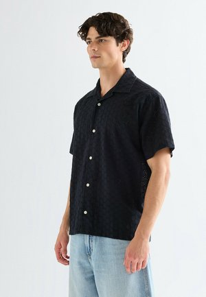 Czarna koszula z krótkim rękawem z teksturowanym heksagonalnym wzorem, z kołnierzykiem i kontrastującymi jasnymi guzikami. Noszona z jasnoniebieskimi dżinsami.