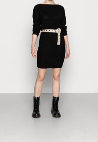 Robe tricotée noire à manches longues, de forme ajustée, avec une ceinture beige ornée de boutons ronds. Associée à des bottes à lacets noires à hauteur de cheville.