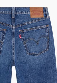Jeans de mezclilla azul oscuro con una parche de cuero marrón con detalles grabados, dos bolsillos traseros con costuras en forma de arco y una pequeña etiqueta roja.
