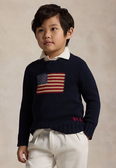 Young boy wearing a navy blue sweater with an American flag design and white pants, standing against a plain beige background.