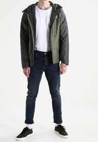 Isolerad jacka i olivgrön och grå med huva, dragkedja framtill och sidofickor, kombinerad med mörka jeans och svarta sneakers.