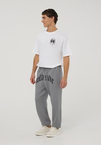 T-shirt bianca a maniche corte con stampa grafica nera sul petto, abbinata a pantaloni della tuta grigi con la scritta "wild side" in grassetto nero.