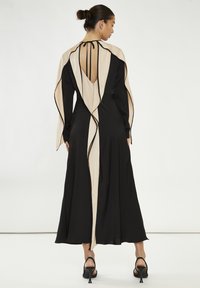 Abito lungo nero con sovrapposizione beige fluente, presenta un profondo scollo a V sulla schiena e piping nero lungo i bordi, realizzato in tessuto morbido.