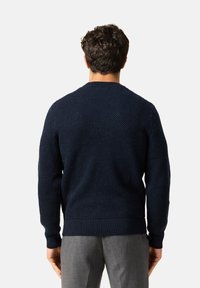 Laivastonsininen neulepaita, jossa on teksturoitu kuvio, pyöreä kaula-aukko sekä resorit hiha- ja vyötäröosassa. Yhdistetty harmaisiin räätälöityihin housuihin.