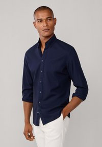 Camicia blu navy con bottoni, maniche lunghe, polsini arrotolati e una texture morbida. Presenta un piccolo logo sul petto e bottoni a contrasto.