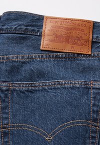 Jeans aus Denim mit einem braunen Lederpatch, das geprägten Text und Bilder zeigt, dunkelblaudem Stoff und kontrastierenden orangefarbenen Nähdetails.