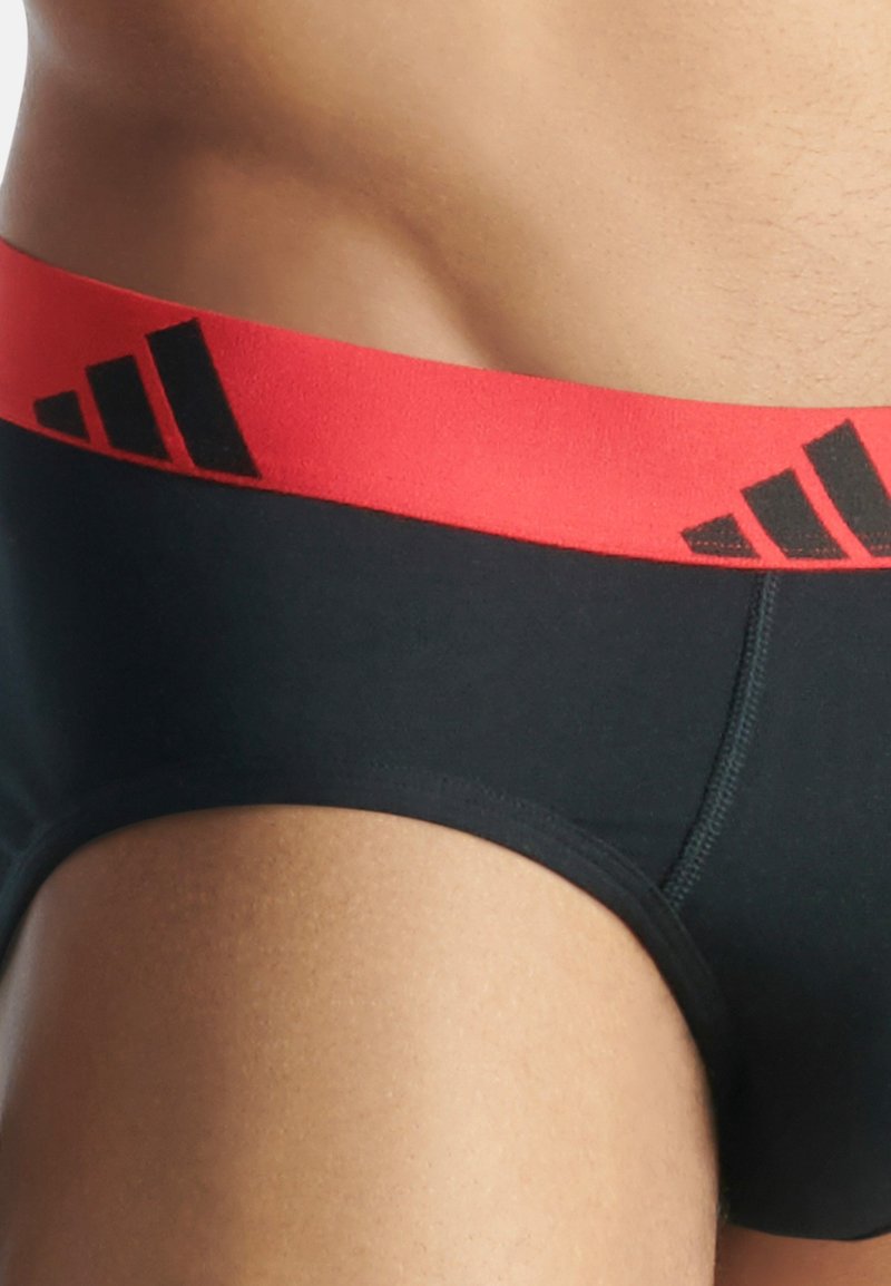 Boxer aderenti neri con vita elastica rossa che presenta un logo nero. Texture liscia, design aderente e cuciture a contrasto lungo le cuciture.