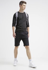 Sort langærmet skjorte parret med sorte shorts, der har en glat tekstur. Hvide sneakers og en grå rygsæk tilføjer kontrast.