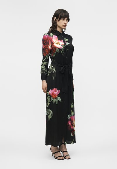 Mujer con un vestido largo de manga larga negro con grandes estampados florales en rosa y rojo, cintura anudada y tacones negros con tiras.
