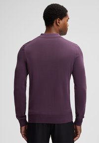 Uomo che indossa un pullover a maniche lunghe viola aderente con colletto, in piedi di spalle contro uno sfondo grigio chiaro uniforme.