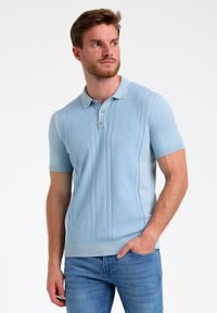Polo de manga corta azul claro con un diseño texturizado, tapeta de tres botones y puños y dobladillo acanalados. Llevado con unos jeans azul claro.