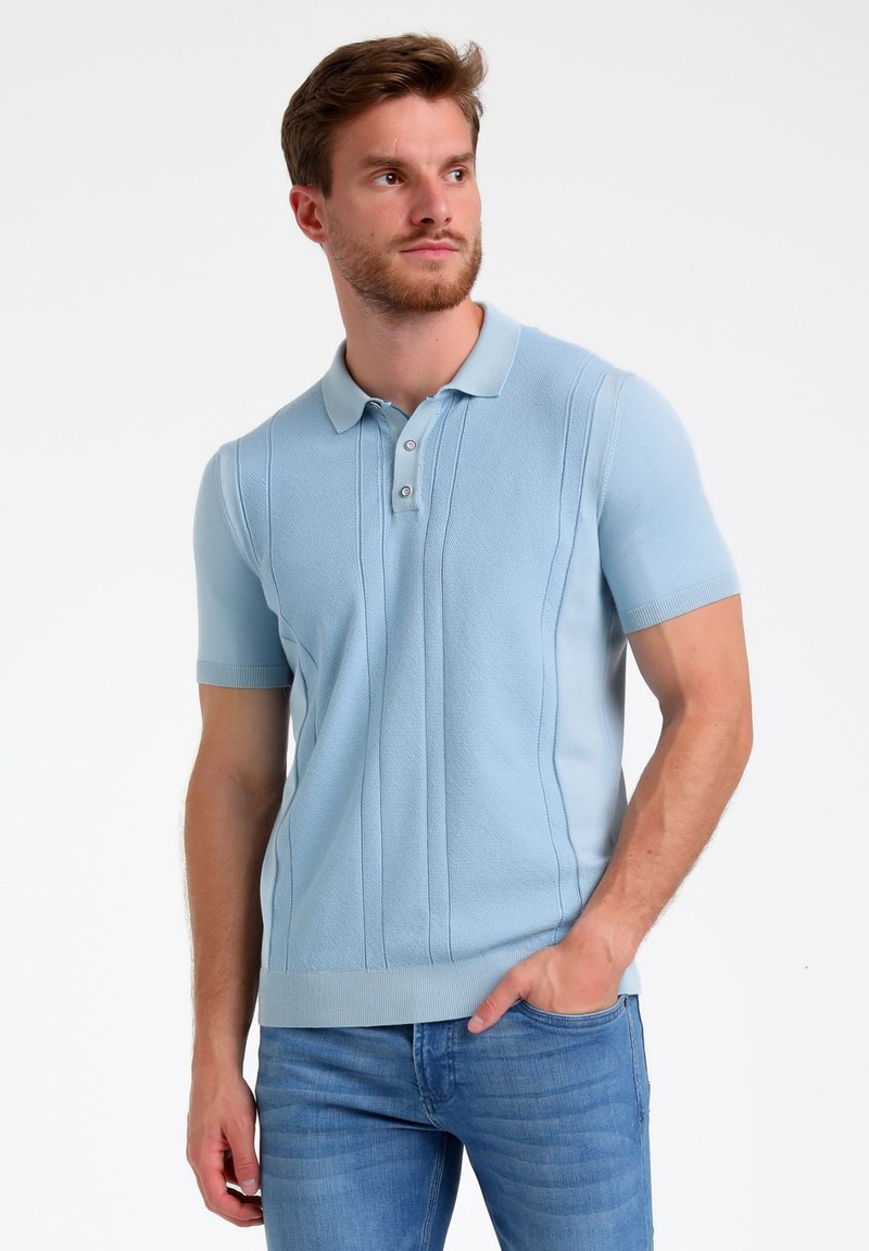 Polo de manga corta azul claro con un diseño texturizado, tapeta de tres botones y puños y dobladillo acanalados. Llevado con unos jeans azul claro.