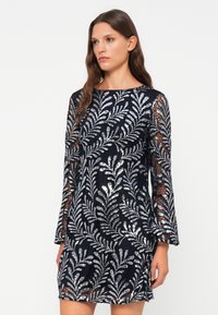 Marinekjole med lange, klokkeformede ermer, med glitrende sølvbladmønstre på en teksturert blondemønster. Kort hemline.