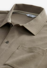 Beige Baumwoll-Polohemd mit Kragen, Brusttasche und schwarzen Knöpfen. Der Stoff hat eine glatte Textur und dezente Nähte.