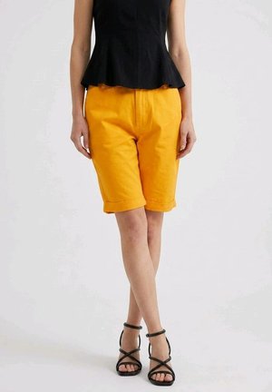 Mujer con top negro sin mangas, pantalones cortos de color amarillo brillante hasta la rodilla y sandalias negras de tacón alto con tiras, de pie con las piernas cruzadas.