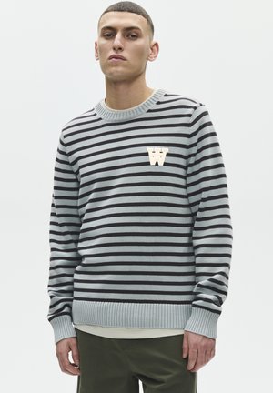 Mann trägt einen grauen und schwarzen gestreiften Pullover mit einem weißen "W"-Emblem auf der Brust, kombiniert mit dunkelgrünen Hosen, stehend vor weißem Hintergrund.