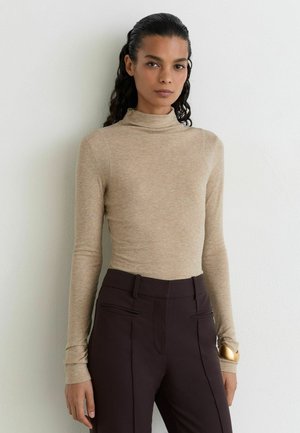 Donna con lunghi capelli ricci che indossa un dolcevita beige e pantaloni marrone scuro a vita alta, in piedi contro un muro bianco.