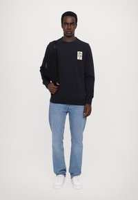 Merimarin pullover kanssa logopatch, yhdistettynä vaaleansinisiin farkkuihin. Malli pitää mustaa olkalaukkua ja on pukeutunut valkoisiin lenkkareihin.