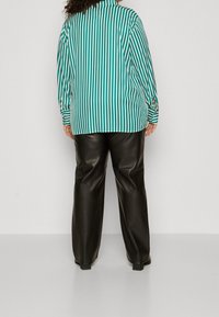 Chemise à rayures vertes et blanches à coupe décontractée, associée à un pantalon en cuir noir. La chemise présente un détail de boutons sur les manches.
