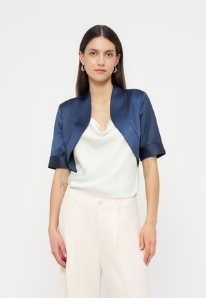 Naine, kellel on pikad tumedad juuksed, kuldsed rõngaskõrvarõngad, tumesinist värvi lühikeste varrukatega bolerojope, valge kaelakaelusega pluus ja kreemjad püksid.