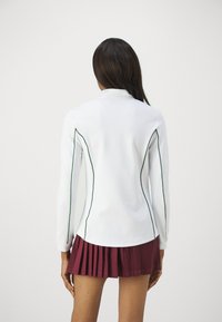 Polo blanco de manga larga con ribetes verdes a los lados, combinado con una falda plisada color burdeos. Textura suave y diseño deportivo.