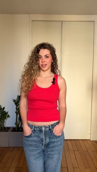 Γυναίκα με σγουρά μαλλιά, που φοράει κόκκινο αμάνικο και μπλε τζιν, στέκεται σε εσωτερικό χώρο με τα χέρια στις τσέπες, φυτά και κλειστές πόρτες πίσω της.