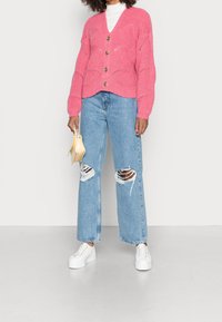 Rosa stickad cardigan med V-hals, prydd med stora guldknappar och ett texturerat mönster. Kombinerad med ljusblå slitna jeans och vita sneakers.