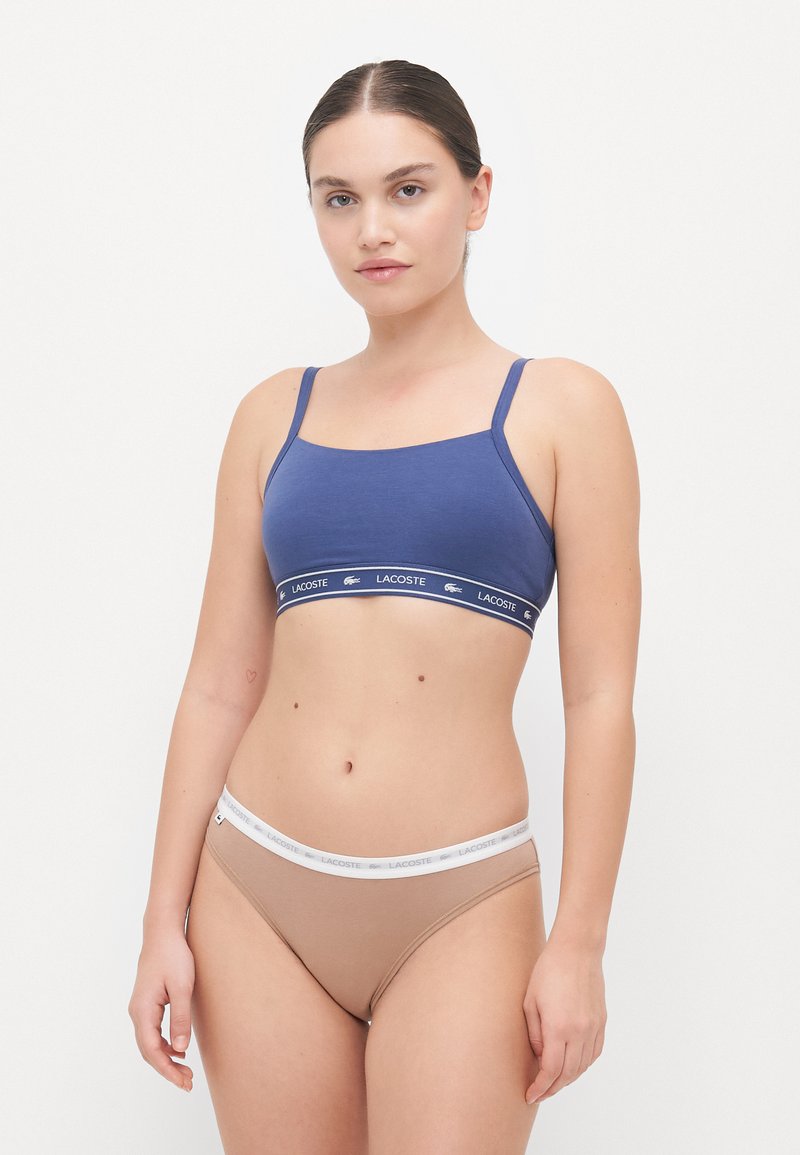 Sötétkék bralette állítható pántokkal, fehér derékszíjjal, amelyen a "LACOSTE" felirat szerepel, bézs alacsony derekú bugyival párosítva.