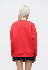 Κόκκινη μπλούζα με χαλαρή γραμμή, διαθέτει στρογγυλή λαιμόκοψη και ριμπ μανσέτες. Μαλακή υφή, χωρίς ορατά μοτίβα ή λεπτομέρειες από μεταλλικά στοιχεία.