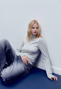 Hellgrauer Rollkragenpullover mit gerippten Bündchen, kombiniert mit faded blauen Oversized-Jeans. Das Model sitzt vor einer weißen Wand auf einem blauen Boden.