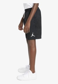 Pantalones cortos negros con cinturilla elástica y cordón blanco, presentando un logo de Jordan en blanco en el lado izquierdo. Combinados con zapatillas blancas.